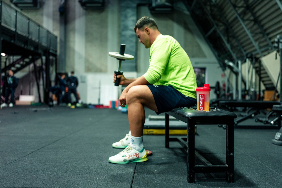 Sportovec odpočívá v posilovně na lavičce, vedle shaker ENERVIT