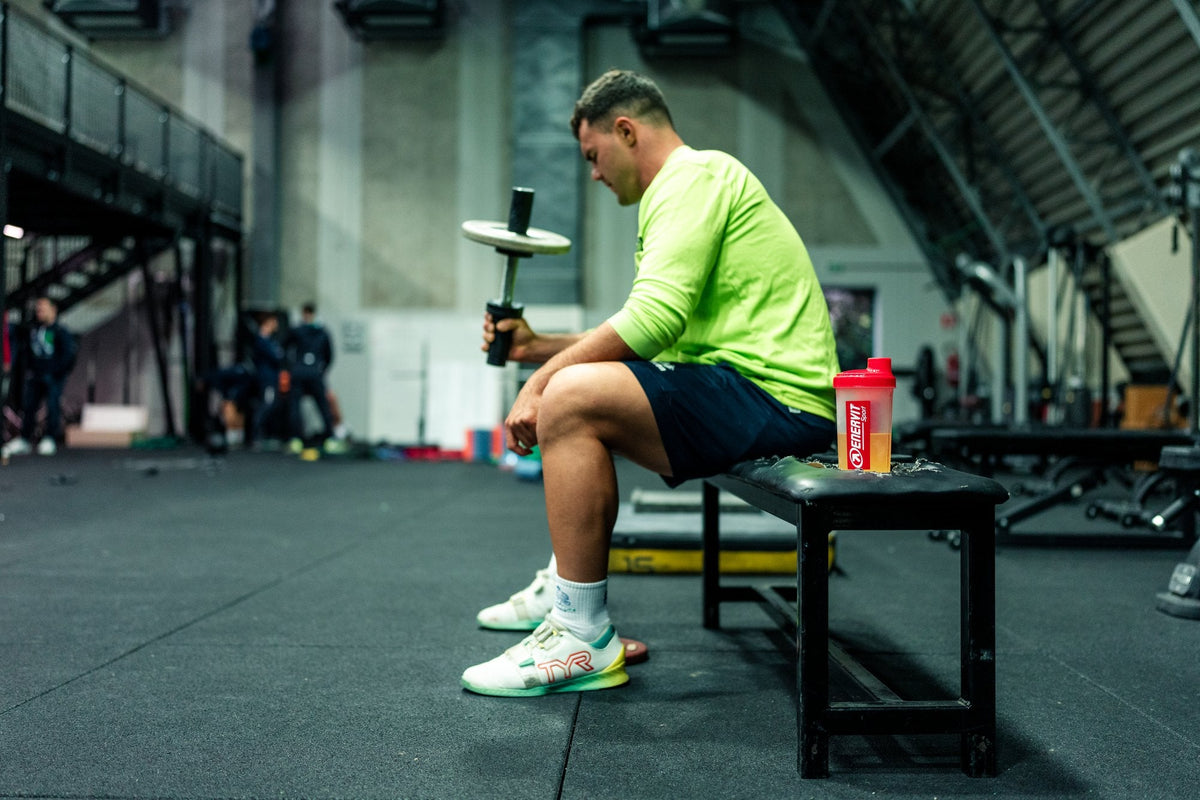 Sportovec odpočívá v posilovně na lavičce, vedle shaker ENERVIT